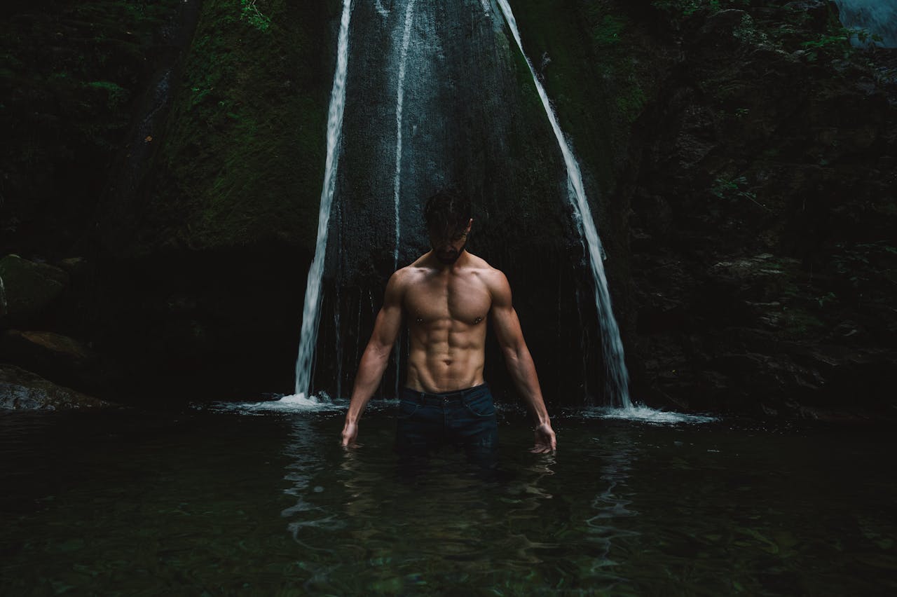 A shirtless man stands in water, showcasing muscles under a mystical waterfall.