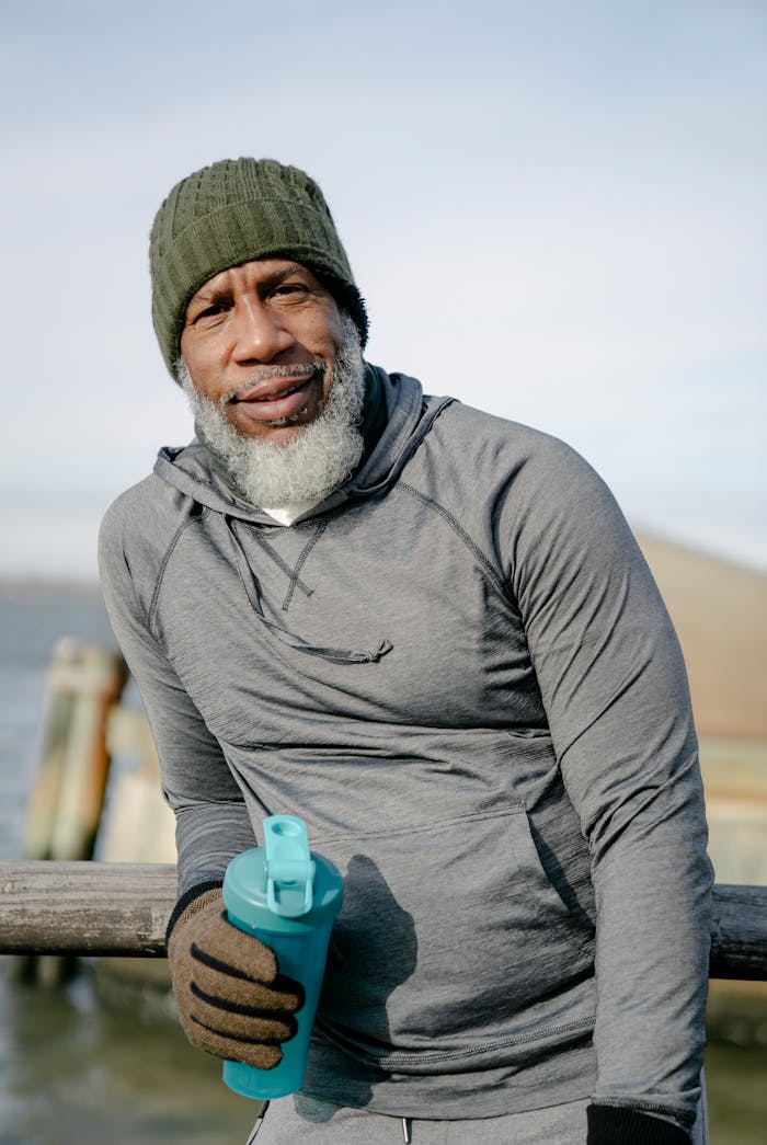 Positive bearded African American male in sportswear and gloves with fitness bottle leaning on fence of bridge